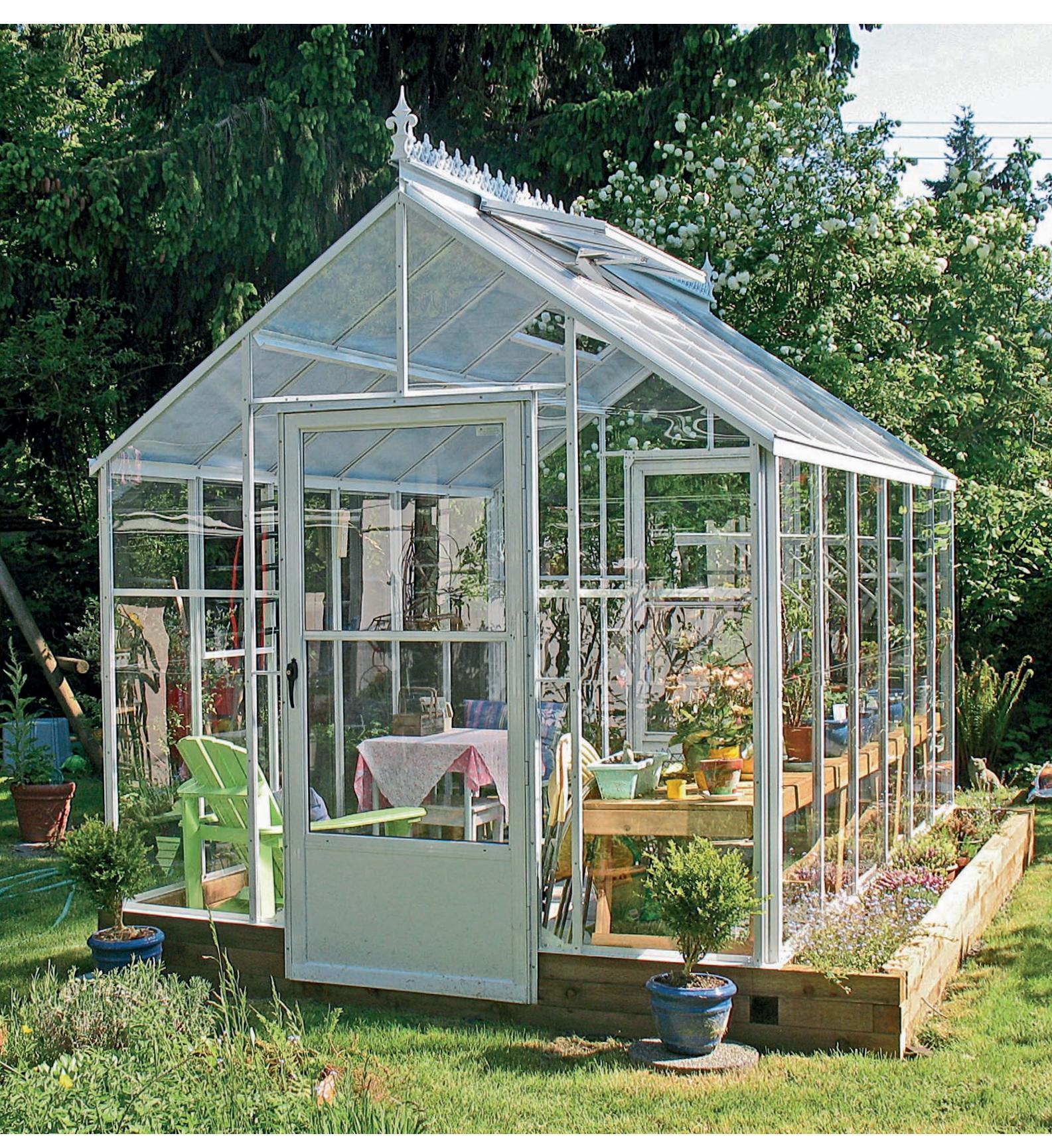 A large Victorian-style glass greenhouse with a drop door, steep roof pitch, and distinctive top crest.